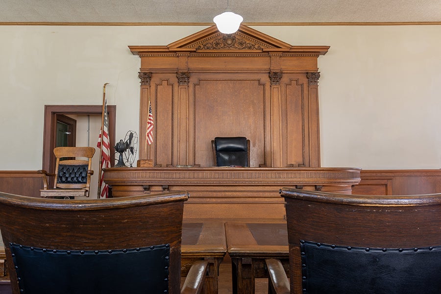 A courtroom interior with a wooden judge’s bench, an empty judge’s chair, and U.S. flags in the background.